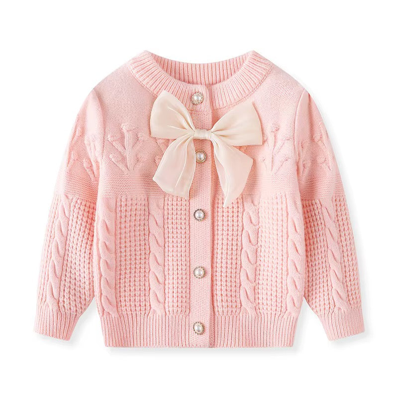 A light pink baby cardigan sweater featuring a large cream-colored satin bow at the neckline, decorative cable knit patterns on the sleeves, and a front button closure with pearl-like buttons.
