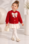 A young girl wearing a red cable-knit cardigan sweater with a large cream bow, a cream pleated skirt, cream tights, and cream shoes, holding a small cream handbag. She is standing in a bright, modern studio setting with white columns and a vase of flowers.